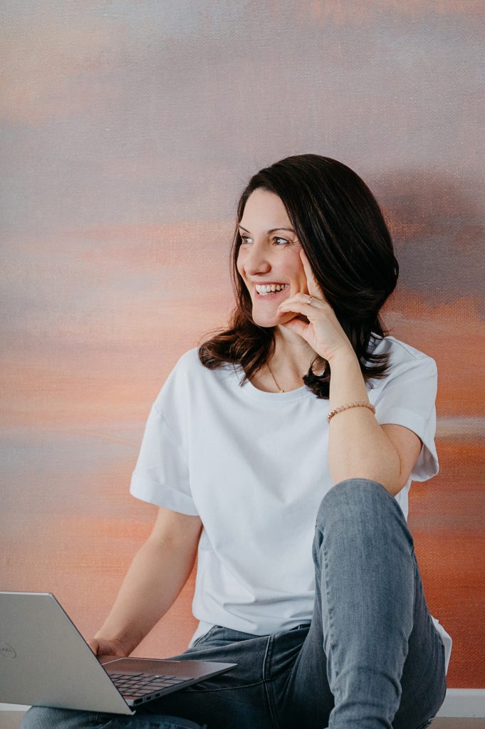 Inhaberin Dana Frey lachend mit Hand abstützend vor einer bunten Wand mit Laptop